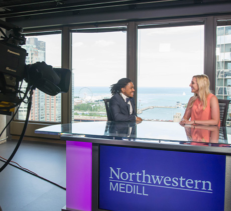 Two students in formal attire talk while seated at a news desk in a tall building with Lake Michigan in the background.