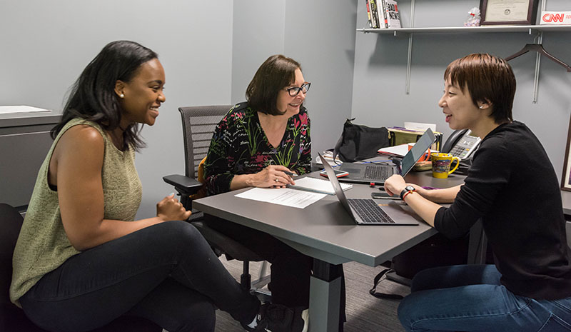 A faculty member speaks with two students who are sitting around a table.