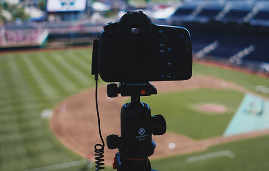 A camera in the foreground points toward a baseball field in the background