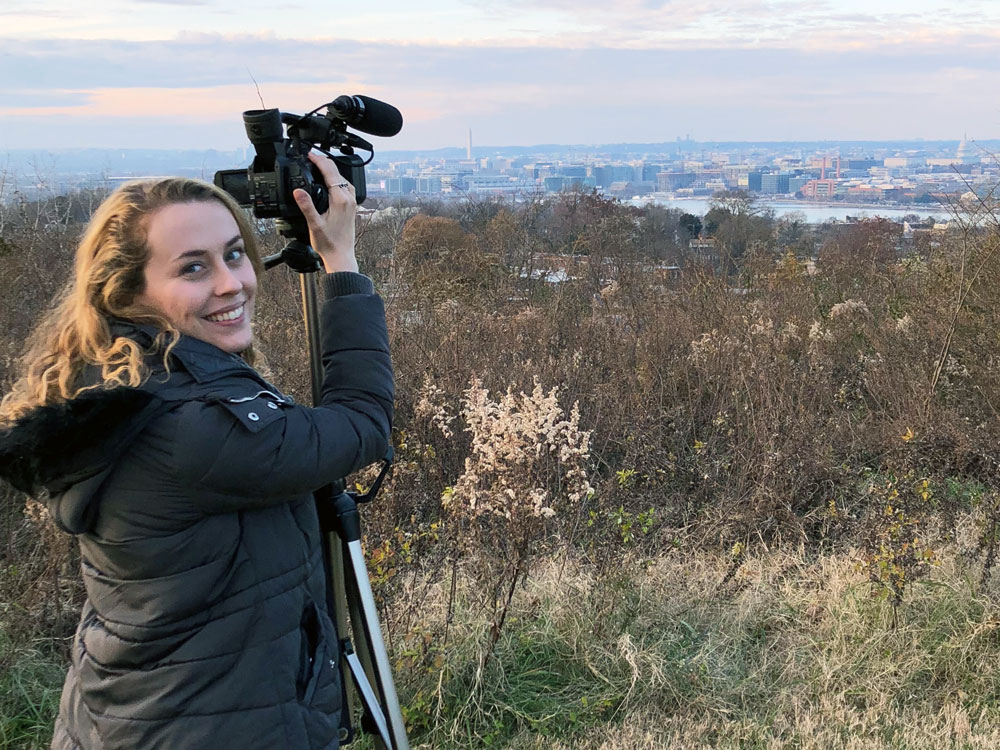 A woman stands far back from a city and adjusts a camera to take a photo of the skyline