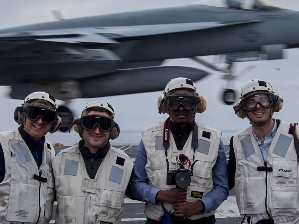 Four people stand wearing helmets, protective glasses and ear protectors onboard a U.S. Naval aircraft carrier