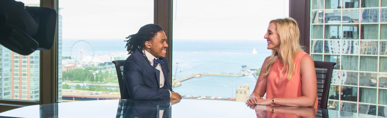 Man and woman smiling at each other as they sit at the Northwestern Medill anchor desk overlooking Lake Michigan. 