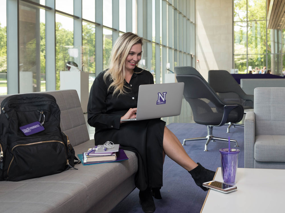A student sits on a couch in a modern-looking lobby space. She works on a laptop computer that has an "N Medill" logo sticker on it.