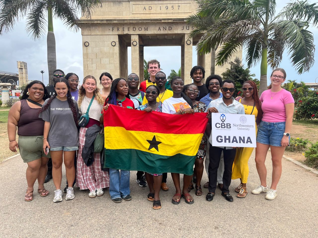 Diverse group of students and staff smiling beneath an arch, holding a Ghana flag and “CBB Northwest Ghana” sign amid tropical palm trees.