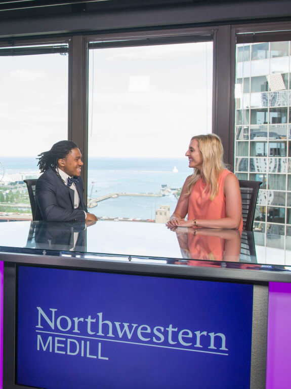 Man and woman smiling at each other as they sit at the Northwestern Medill anchor desk overlooking Lake Michigan. 