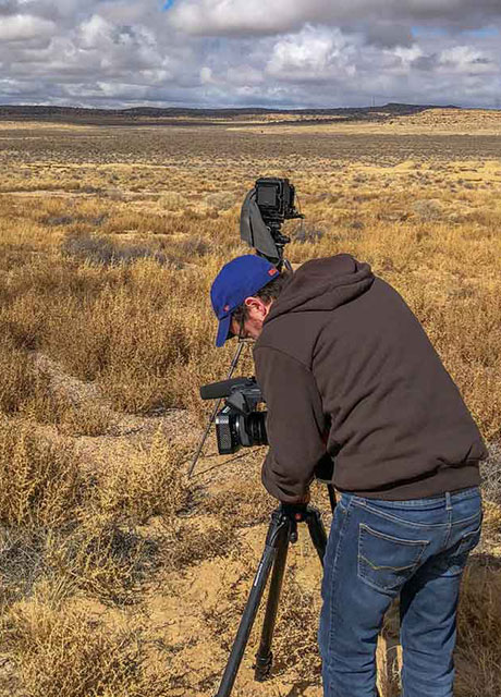 A man standing behind a tripod and camera looking out at a vast field.