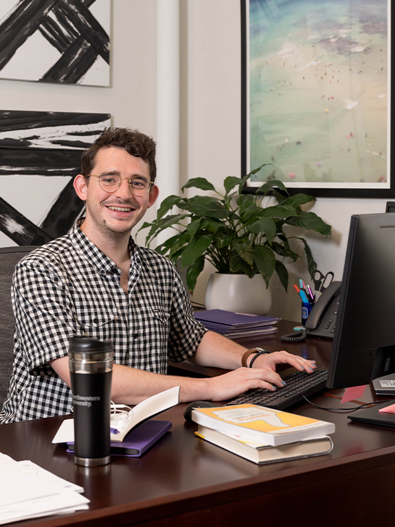 A working professional sits at a computer in an office with books on the desk