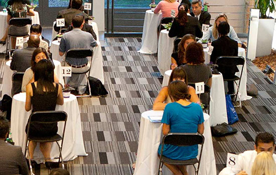 Groups of two sitting at many tables having interviews in a big room.