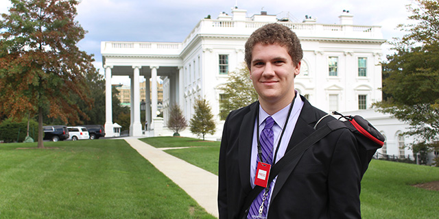 Student wearing a suit and carrying camera equipment in front of White House.