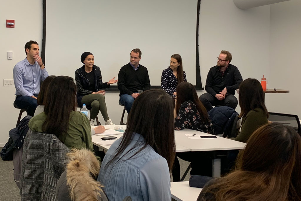 A panel of five people sit on chairs at the front of a room and speak to a group of students who are seated at tables