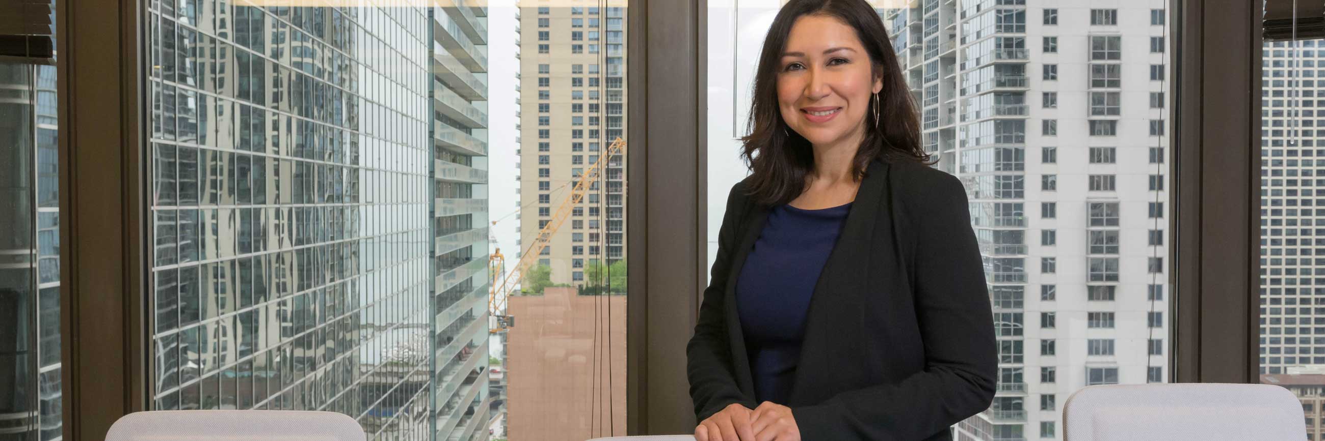 A woman in a suit coat stands by a conference table. There are high-rise buildings in downtown Chicago outside the window.