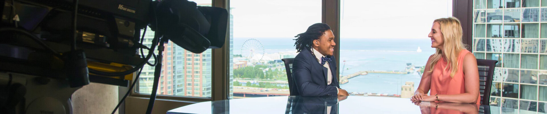 Man and woman smiling at each other as they sit at the Northwestern Medill anchor desk overlooking Lake Michigan. 