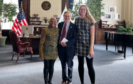 A professor and two students smile and pose in a formal office beside American and state flags, with a grand piano, bookshelves, and framed photos behind them.