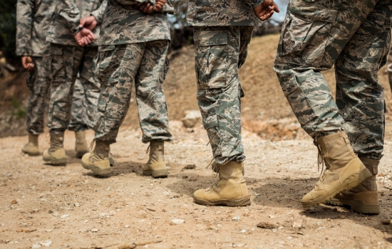 Group of soldiers in camouflage uniforms and tan combat boots walk forward across a dusty, rocky trail.