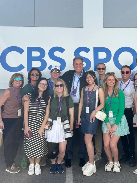 Smiling group of students poses outside a CBS Sports bus, wearing event badges.