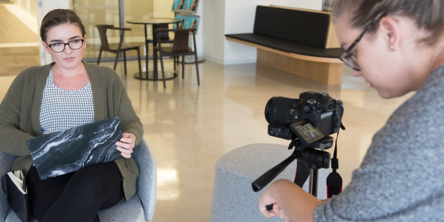 Woman holds a laptop during a filmed interview, while another person operates a camera on a tripod in a bright room.