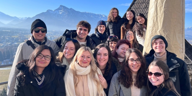Smiling group of students pose at a mountain lookout, with snowy peaks and a bright blue sky behind them.