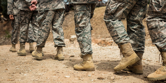 Close-up of soldiers in camouflage uniforms and tan combat boots marching across a dusty dirt road.