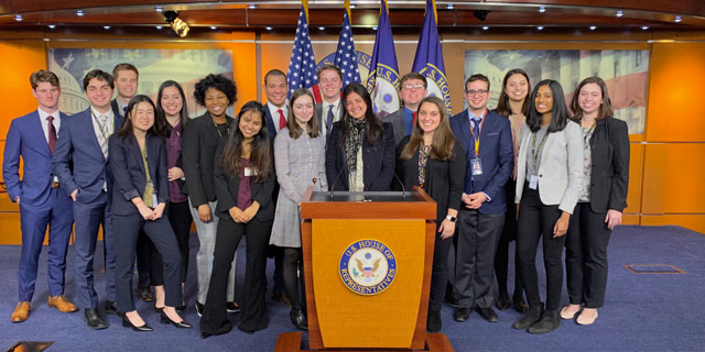 Diverse group of students pose smiling behind a podium bearing a state seal, with flags in press briefing room.