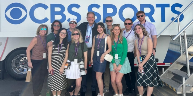 Smiling group of students poses outside a CBS Sports bus, wearing event badges.