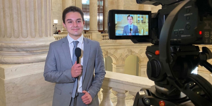 Student reporter in a gray suit holds a microphone inside the Rotunda, while a professional video camera records him in the foreground.