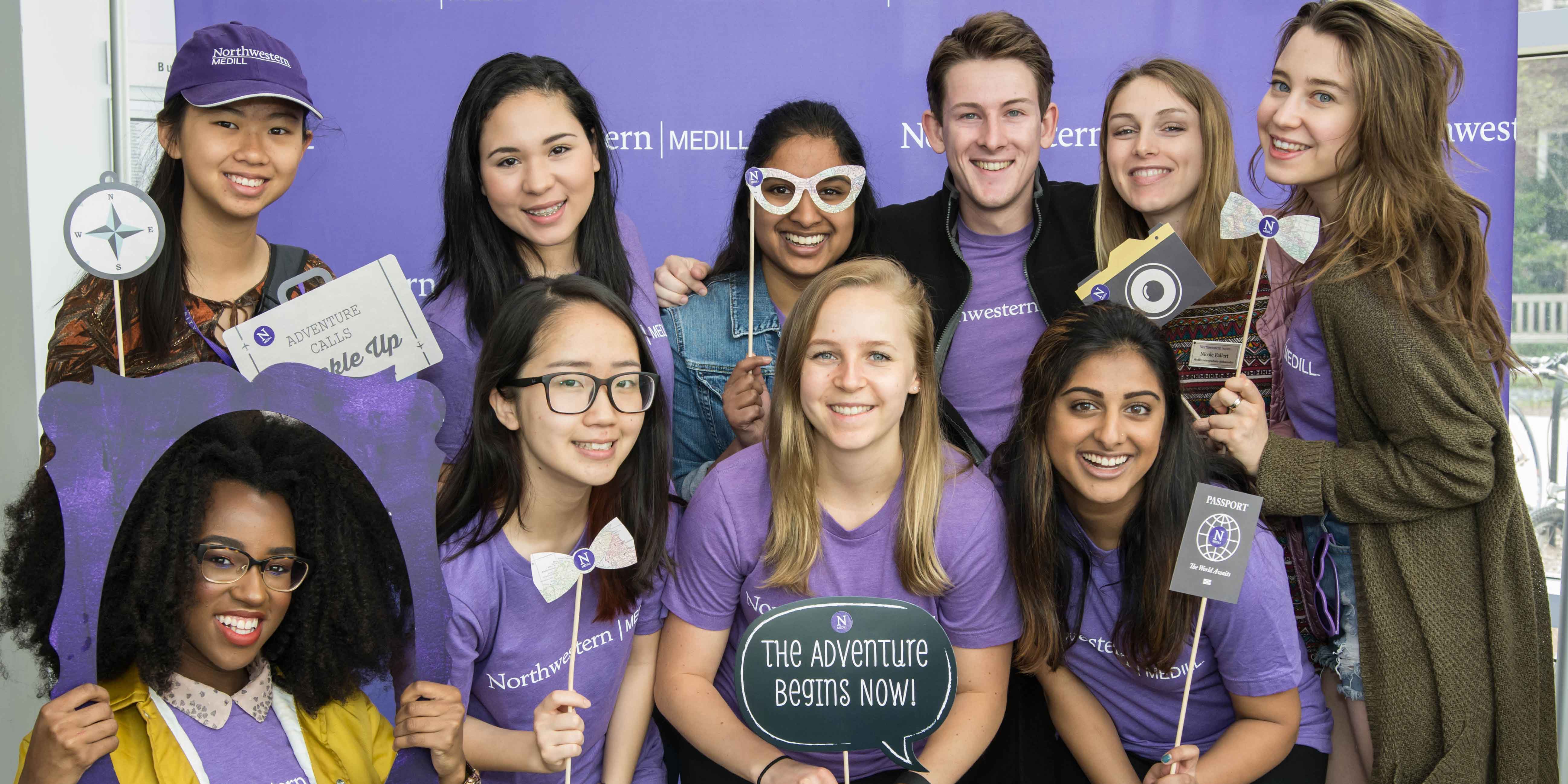 Students wearing Medill shirts holding props and posing in front of a purple photo background. 