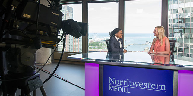 Two students in formal attire talk while seated at a news desk in a tall building with Lake Michigan in the background.