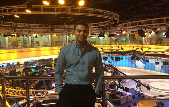 Student standing and smiling in front of a newsroom.