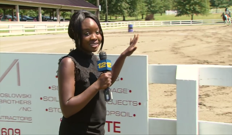 Student standing outside talking into a microphone. There's a fence and a dirt racetrack in the background.