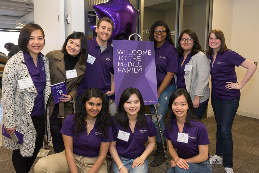 A group of students wearing purple shirts with the Northwestern Medill logo on them stand by a sign that says "Welcome to the Medill family".