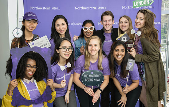 Students wearing Medill shirts smile with props in front of a Medill photo booth.