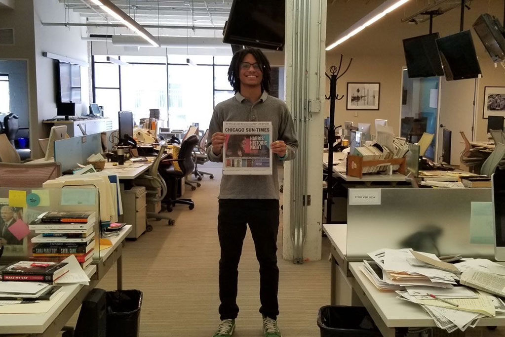 Adam Mahoney stands in the Chicago Sun-Times office smiling and holding a copy of the newspaper.
