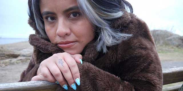 A woman wearing a fur coat looks into the camera while seated on a bench facing a body of water.