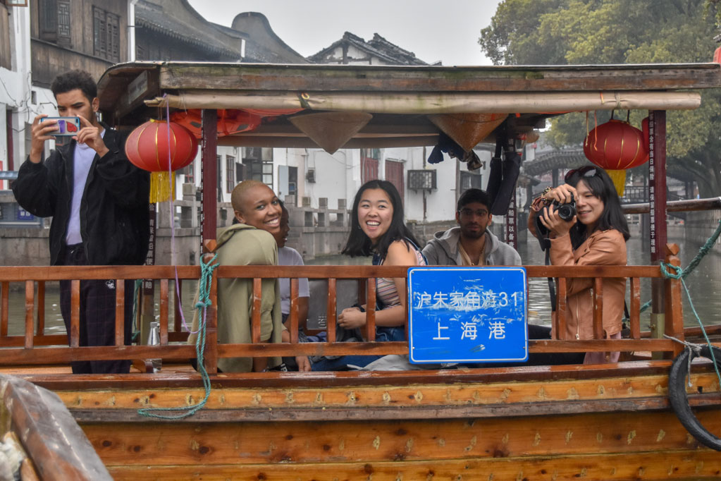 Students take photos while riding on a boat with buildings in the background.