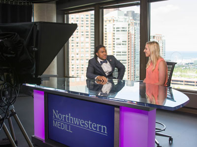 A student interviews a guest at a broadcast desk with the Chicago skyline in the background.