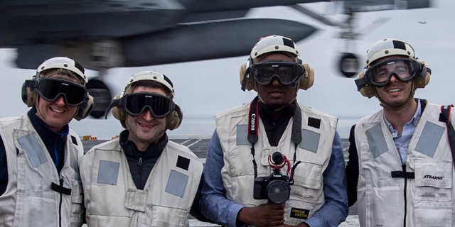 Four people stand on a Naval carrier wearing helmets and protective glasses and headphones.