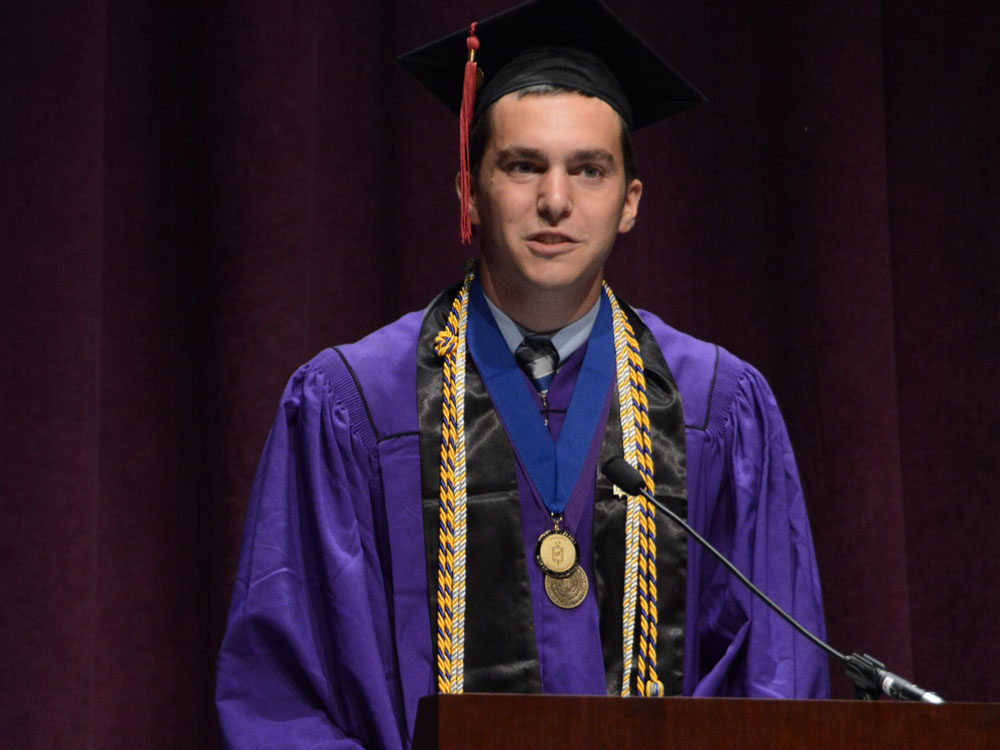 A male student stands behind a podium and speaks; he is wearing a purple graduation cap and gown
