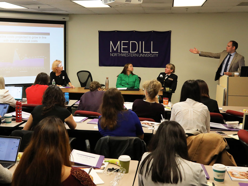 A panel of experts sits at a table at the front of a room and a man who's standing speaks to the group of students in the room