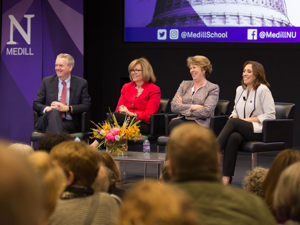 Three women and one man sit on stage during a panel discussion and smile at the crowd