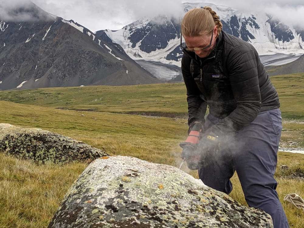 Jess Stevens, an environmental science teacher at Gary Comer College Prep High School in Chicago, drills into a deposited boulder in the valley of the Potanin Glacier in Mongolia. The rock chipping she gathers could hold the clue as to how fast the climate is warming.