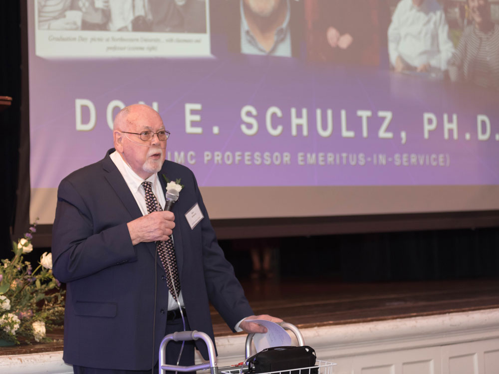 Don Schultz stands at the front of a room and speaks into a microphone at the Hall of Achievement reception