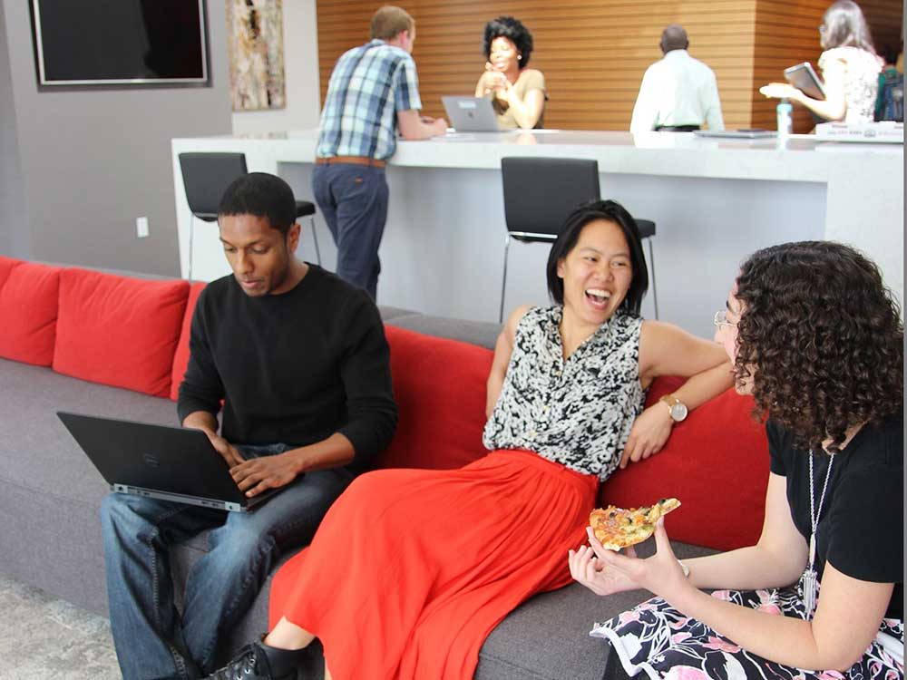 Two women talk and laugh on a couch while a man works beside them on a laptop.