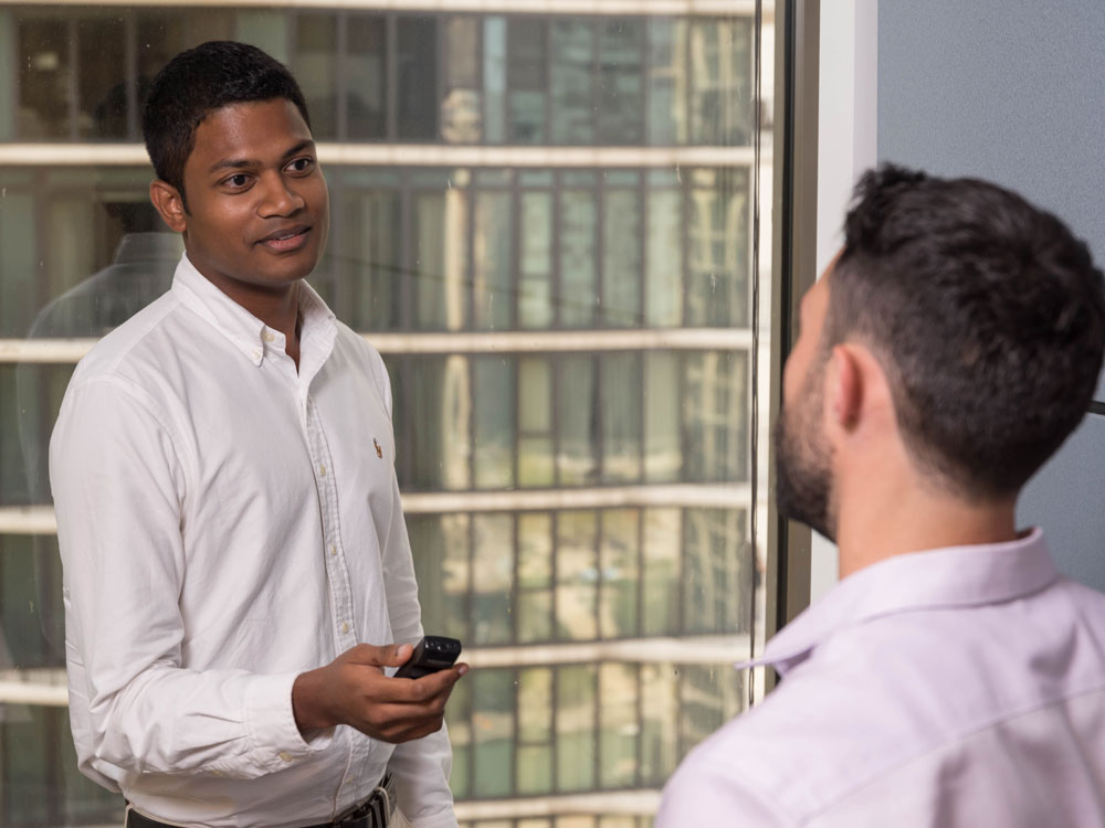 A student holds a recording device while he interviews someone. A high-rise building can be seen out the window.