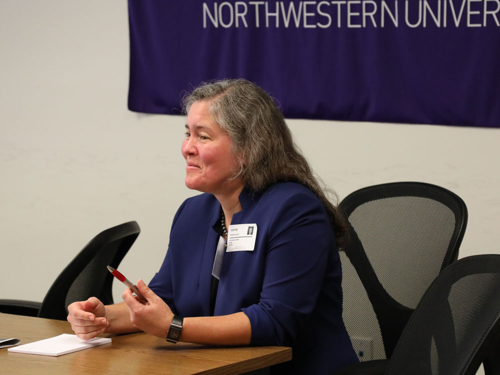 A woman sits at a table at the front of a room and talks to a crowd