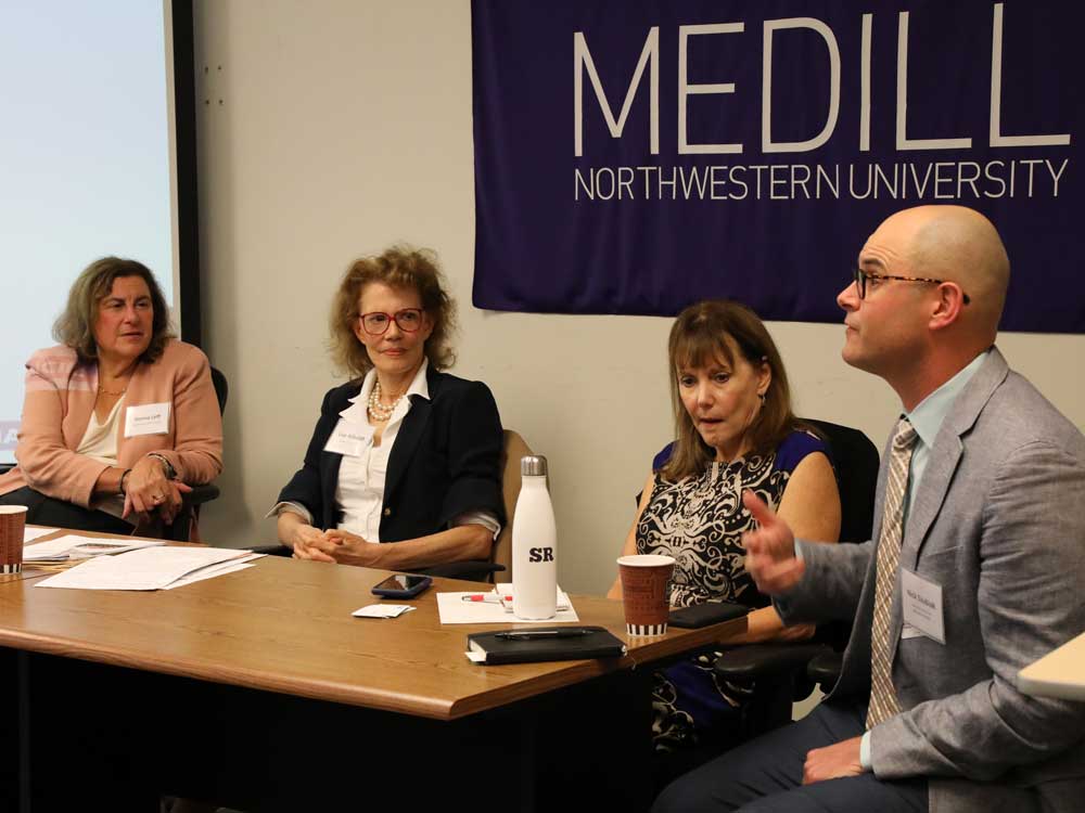 Four people sit at a table in the front of a room; one person is talking.