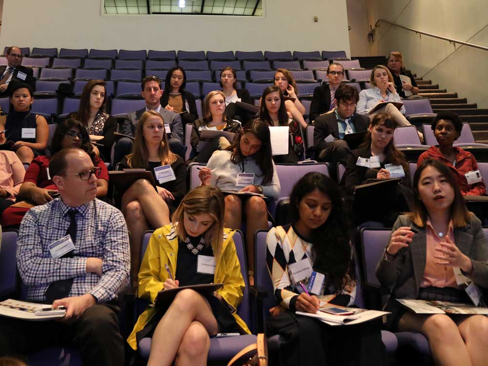 Students sit in auditorium seats. Some students are taking notes. One student is asking a question.