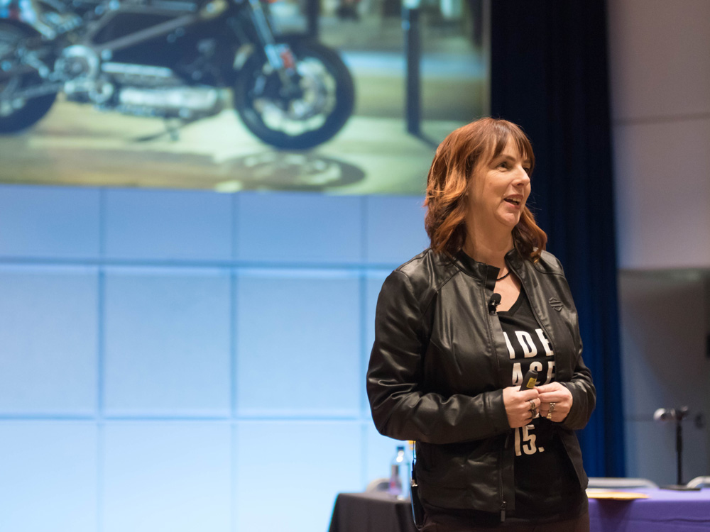 A woman stands on stage and speaks. A motorcycle is pictured on the screen behind her.