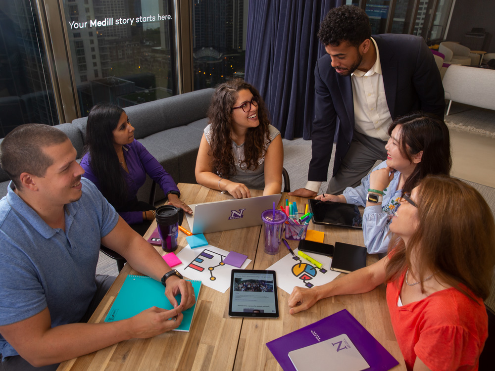 A group of people sit around a table and talk, computers and notebooks are on the table