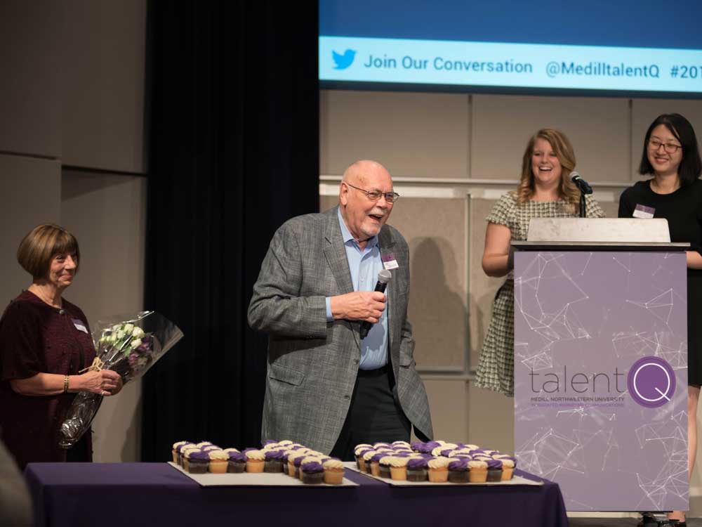 A man holds a microphone and speaks to a crowd; a table filled with cupcakes sits before him