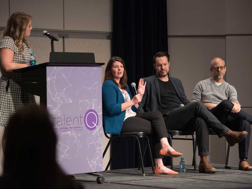 Three panelists sit in chairs on stage and speak to a crowd